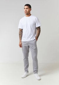 Valge puuvillane lühikeste varrukatega t-särk, kombineeritud heledatehallide pükste ja valgete tossudega. Modell seisab käed taskutes.