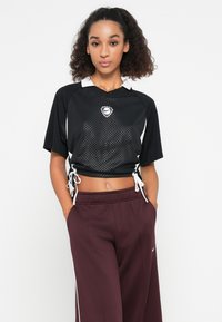 Juodas trumpas sportinis marškinėlis su tinklinio tekstūra, baltu apykakle ir virvutėmis šonuose. Sujungtas su bordo spalvos sportinėmis kelnėmis, turinčiomis baltus akcentus.