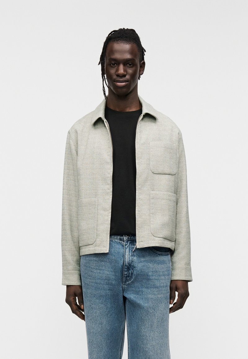 Veste gris clair texturée avec un col, deux poches avant et une fermeture éclair, portée sur une chemise noire et un jean bleu.