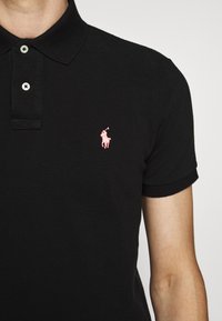 Polo noir en tissu texturé avec une coupe ajustée, deux boutons et un logo brodé rose sur la poitrine gauche.