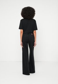 Άτομο με σγουρά μαλλιά στέκεται πλάτη φορώντας μαύρο κοντομάνικο πουκάμισο και μαύρο τζιν παντελόνι με ψηλή μέση και φαρδιά μπατζάκια σε λευκό φόντο.