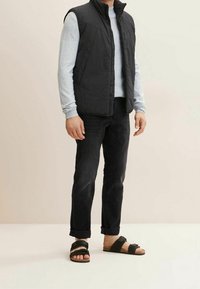 Chaleco negro sin mangas sobre una camisa de manga larga gris claro, combinado con jeans negros y sandalias negras de doble tira. Texturas suaves.