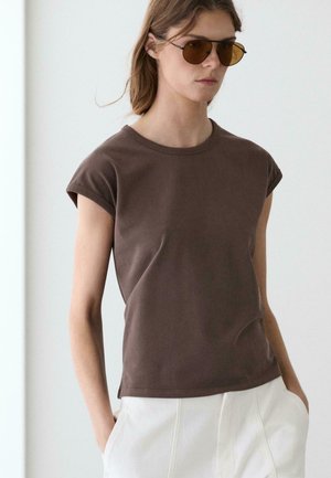 Donna con indosso una maglietta marrone a maniche corte, pantaloni bianchi e occhiali da sole con lenti ambra, in piedi davanti a uno sfondo chiaro e uniforme.