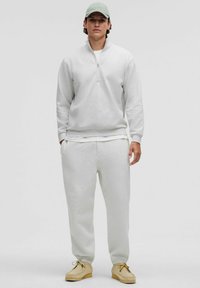Helle graue Zip-Up-Sweatjacke mit hohem Kragen, kombiniert mit passenden Jogginghosen. Aus weichem, strukturiertem Stoff und in lockerer Passform. Braune Schuhe.