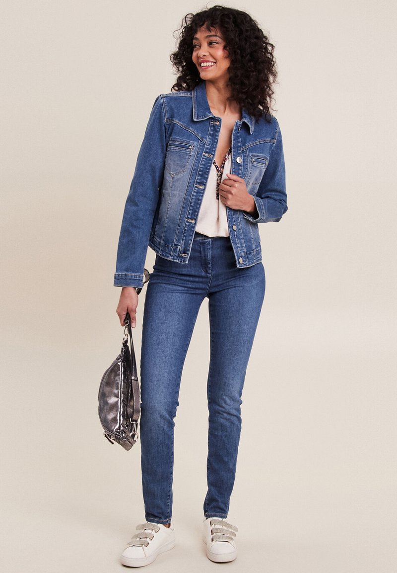 Veste en denim bleu moyen, avec des poches avant et une fermeture à boutons. Associée à un jean skinny bleu et des baskets blanches. Tient un sac argenté.