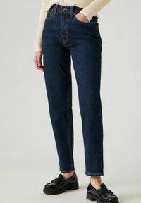 Donkerblauwe denim jeans met een hoge taille, rechte pasvorm, contrasterende stiksels en traditionele vijf-pocketontwerp. Gestyled met zwarte loafers.