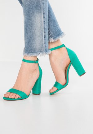 Zielone zamszowe sandały na wysokim obcasie typu słupek, z odkrytym noskiem i paskiem wokół kostki. Charakteryzują się gładką fakturą i minimalistycznym designem.
