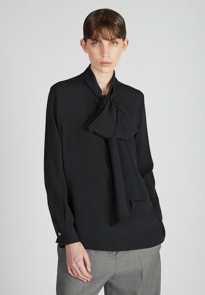 Blusa nera realizzata in tessuto leggero, con un ampio colletto a fiocco, maniche lunghe e polsini con bottoni. Indossata con pantaloni grigi.