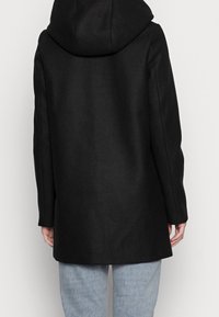 Abrigo de lana negro con capucha, con un corte entallado y diseño recto, visto desde atrás, llevado sobre jeans azul claro.