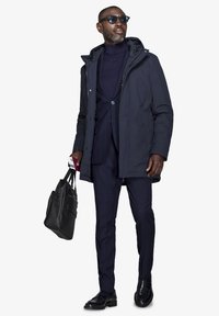Marine vandtæt parka over et tætsiddende marineblåt stribet jakkesæt, sorte lædersko og en sort tote-bag. Inkluderer solbriller og et dokument i hånden.