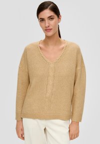 Beige gestrickter Pullover mit V-Ausschnitt, der eine Zopfstrickdetails in der Mitte und gerippte Bündchen aufweist, kombiniert mit cremefarbenen gerippten Hosen.