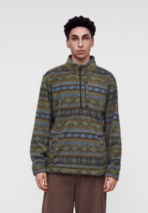 Jeune homme portant un pull en polaire à motifs avec une demi-fermeture éclair et une poche avant, debout devant un fond gris clair uni.