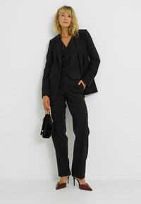 Traje negro a medida con blazer entallado y pantalones, textura suave. Combinado con tacones marrones puntiagudos y un pequeño bolso negro.