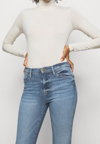 Top de cuello alto de manga larga color beige y jeans azul de lavado medio. Los jeans tienen bolsillos delanteros, cierre de cremallera y un leve detalle desgastado.