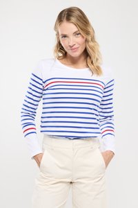 Vrouw met blond haar in een witte langemouwen shirt met blauwe en rode horizontale strepen en crèmekleurige broek, handen in de zakken, tegen een witte achtergrond.