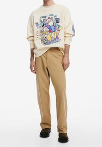 Personne portant un sweat-shirt graphique beige avec un personnage de dessin animé et un pantalon marron clair, debout, les bras détendus, sur un fond blanc uni.