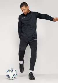 Schwarzes Sporttrikot mit halbem Reißverschluss und weißen Akzenten, kombiniert mit schwarzen, schmal zulaufenden Hosen. Schwarze Fußballschuhe und ein gemusterter Fußball auf dem Boden.