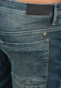 Shorts en denim bleu avec une surface texturée, présentant une patch en cuir noir étiquetée "BLEND" et un bouton en métal à la taille.