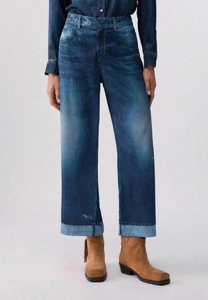 Jeans di denim a gamba larga e taglio corto con orli risvoltati e dettagli sbiaditi, abbinati a stivaletti in pelle scamosciata color beige su una persona che indossa una camicia di denim scuro.