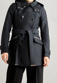 Trench-coat noir imperméable avec un design à double boutonnage, grand col, ceinture et poches zippées avec des accents métalliques et une texture lisse.