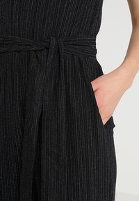 Černý overal s vertikálními pruhy, texturovaná tkanina, páskový úvaz, a boční kapsy. Má přiléhavý vršek a splývavou spodní část.