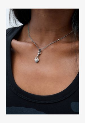 Srebrna ogrlica s malim privjeskom u obliku srca i plamena, smještena na području ključne kosti osobe u crnoj majici.