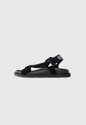 Black sandals with a flat sole, suede strap design, and adjustable ankle strap featuring a white label. Lightweight and minimalistic style.