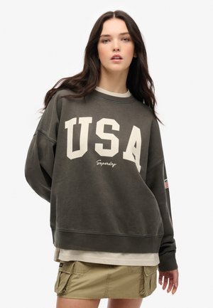 Sweatshirt vert olive avec "USA" en crème, col rond et poignets côtelés. Porté par-dessus une chemise beige clair ; présente un petit patch de drapeau sur la manche.