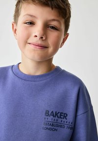 Lilla sviiter, millel on ümar kaelus, ja millel on must logo prinditud vasakul rinnal: "BAKER by TED BAKER ESTABLISHED 1988 LONDON."