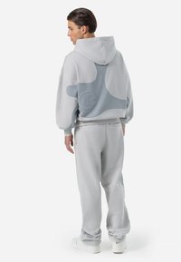 Grijze hoodie met een gestructureerde blauwe accent op de achterkant, vergezeld van bijpassende grijze trainingsbroek en witte sneakers. Casual, relaxte pasvorm.