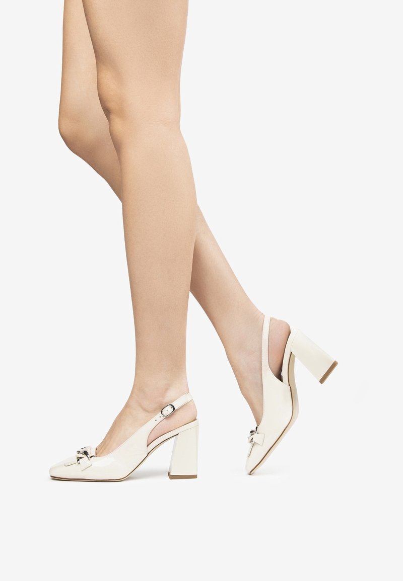 Escarpins blancs en cuir verni avec bride arrière, bout carré, détail nœud et talon bloc épais. Texture lisse avec une finition brillante.