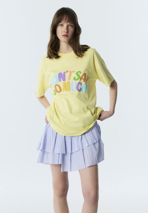 Jeune femme aux cheveux bruns porte un t-shirt jaune avec le texte coloré « NE DIS PAS TROP » et une jupe rayée en couches bleue et blanche.