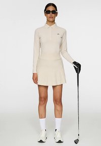 Beżowa koszulka polo z długim rękawem z logo oraz plisowana spódnica. W zestawie z czarnymi okularami przeciwsłonecznymi, białymi skarpetkami i białymi tenisówkami, trzyma kij golfowy.
