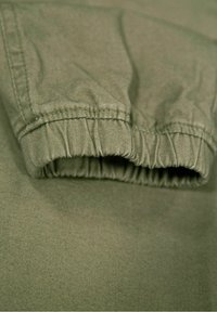 Groene stof met een geplooide manchet detail, die textuurnaad en een gladde oppervlakte toont. De kleur is een gedempt olijfgroen.
