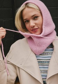 Lyserød ribbet strik hætte med bred hals, båret med en beige frakke over en mønstret sweater. Har en snor ved halsen og tekstureret stof.
