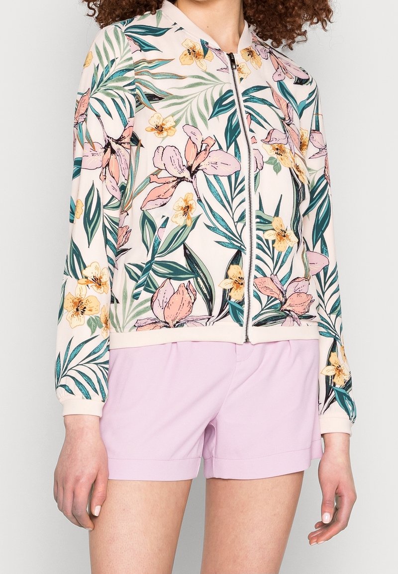 Femme portant une veste zippée à fleurs avec de grandes fleurs roses et jaunes ainsi que des feuilles vertes, associée à un short violet clair.