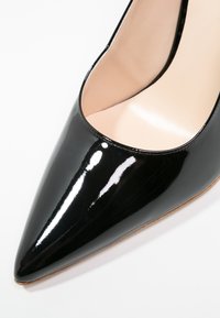 Escarpin en cuir verni noir avec un bout pointu et une finition lisse et brillante. L'intérieur est de couleur claire et la semelle est beige.