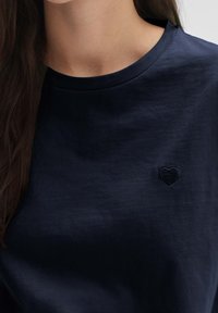 T-shirt in cotone blu navy con collo rotondo e un piccolo ricamo a forma di cuore nero sul lato sinistro del petto. Tessuto morbido.