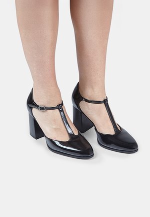 Black patent leather high-heeled shoes with a T-strap design, square toe, and chunky heel. Features cut-out sides and an adjustable buckle.