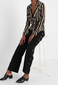 Blazer rayé noir et beige avec un col à volants, associé à un pantalon large noir orné de boutons dorés sur les côtés.