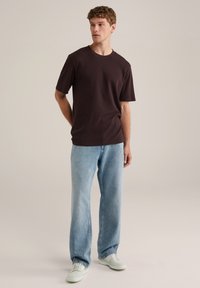 Camiseta de manga corta de color marrón oscuro combinada con jeans rectos de color azul claro y zapatillas blancas, ambientada en un fondo neutral.