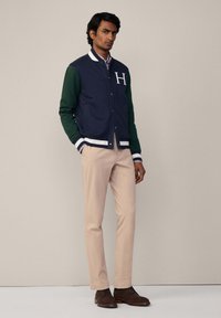 Marineblauwe en groene varsityjack met witte ribgebreide manchetten en kraag, gedragen over een geruite blouse; gecombineerd met lichtbeige broek en bruine schoenen.