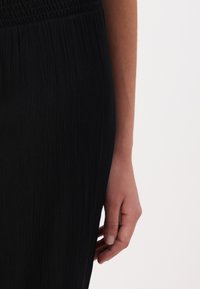 Robe noire texturée avec une taille froncée et un gros plan d'une main touchant le tissu. Le matériau a un aspect froissé.