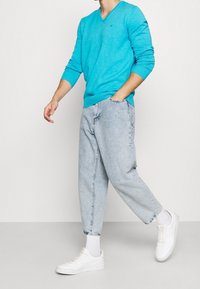 Pull turquoise à col en V, jean bleu clair ample, et baskets blanches avec chaussettes blanches. Look décontracté avec une coupe ample et des détails d'accentuation minimalistes.