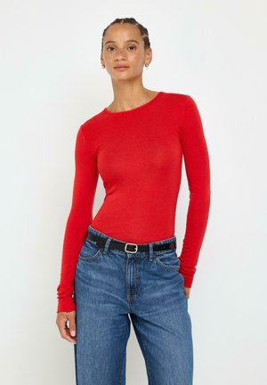 Ung kvinde med fletninger iført en tætsiddende rød langærmet top, blå jeans og et sort bælte mod en hvid baggrund.