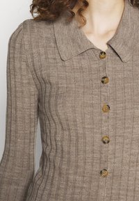 Brun ribbad stickad cardigan med knappstängning framtill, som har en krage och stora bruna knappar. Texturen är mjuk och strukturerad.