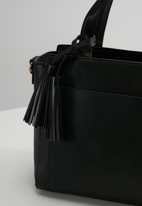 Sac à main en cuir noir avec un design structuré, une texture lisse et un accent décoratif en franges sur le côté. Ferrures dorées visibles.