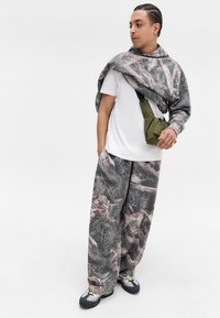 Grå camouflage sweatshirt lagt over en hvid T-shirt, parret med løse grå mønstrede bukser og en grøn posetaske. Sorte og grå sneakers.