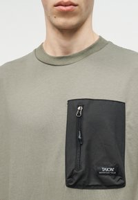 Νεαρό άτομο που φοράει ελιά πράσινο πουκάμισο με μεγάλη μαύρη τσέπη με φερμουάρ, επισημασμένη με την ένδειξη «TAION Mountain Wear» στο στήθος.