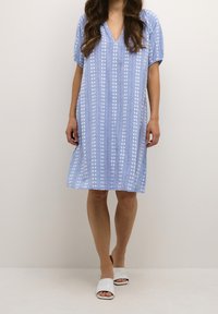 Kvinna i en ljusblå knälång klänning med vita blommiga ränder och vita öppna mule-sandaler, stående mot en enfärgad bakgrund.